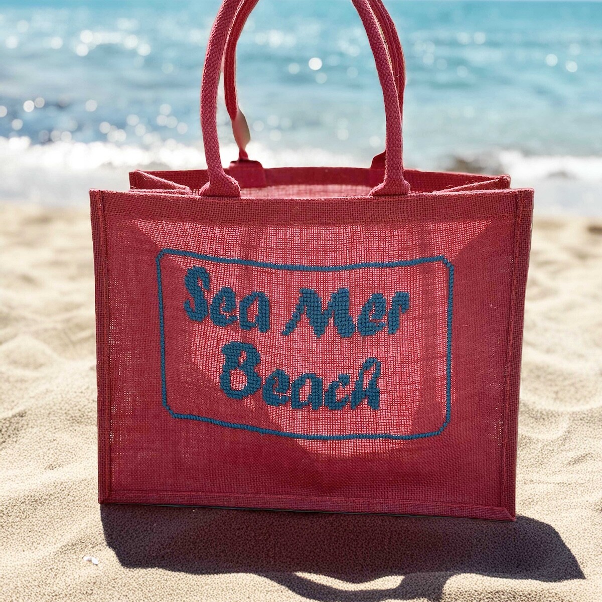 Cabas rouge brodé ‘SEA MER BEACH’ posé sur fond sable clair